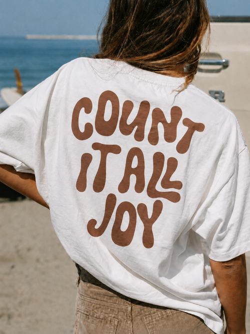 A person viewed from behind wearing a white oversized T-shirt with large brown bubble lettering that reads 'COUNT IT ALL JOY,' standing outdoors near the beach.