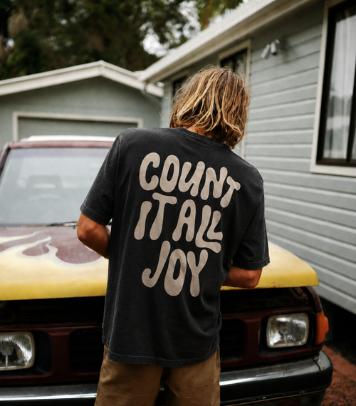 A person standing with their back to the camera in front of a yellow car and a house, wearing a faded black t-shirt with 'COUNT IT ALL JOY' printed in beige on the back.