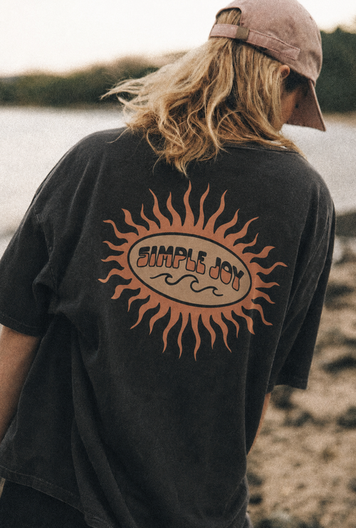 A person viewed from behind wearing a faded black t-shirt with a large orange sun graphic and the words 'SIMPLE JOY' across the back, along with a dusty pink cap, set against an outdoor beach background.