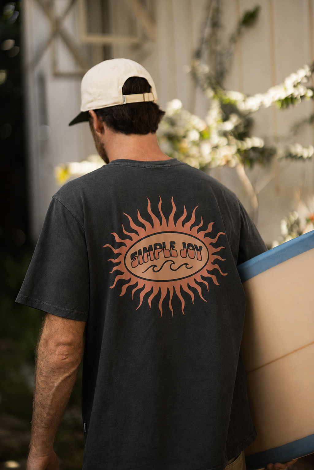 A man viewed from behind wearing a charcoal gray t-shirt with a large orange sun graphic and 'Simple Joy' logo on the back, holding a surfboard against a blurred outdoor background.