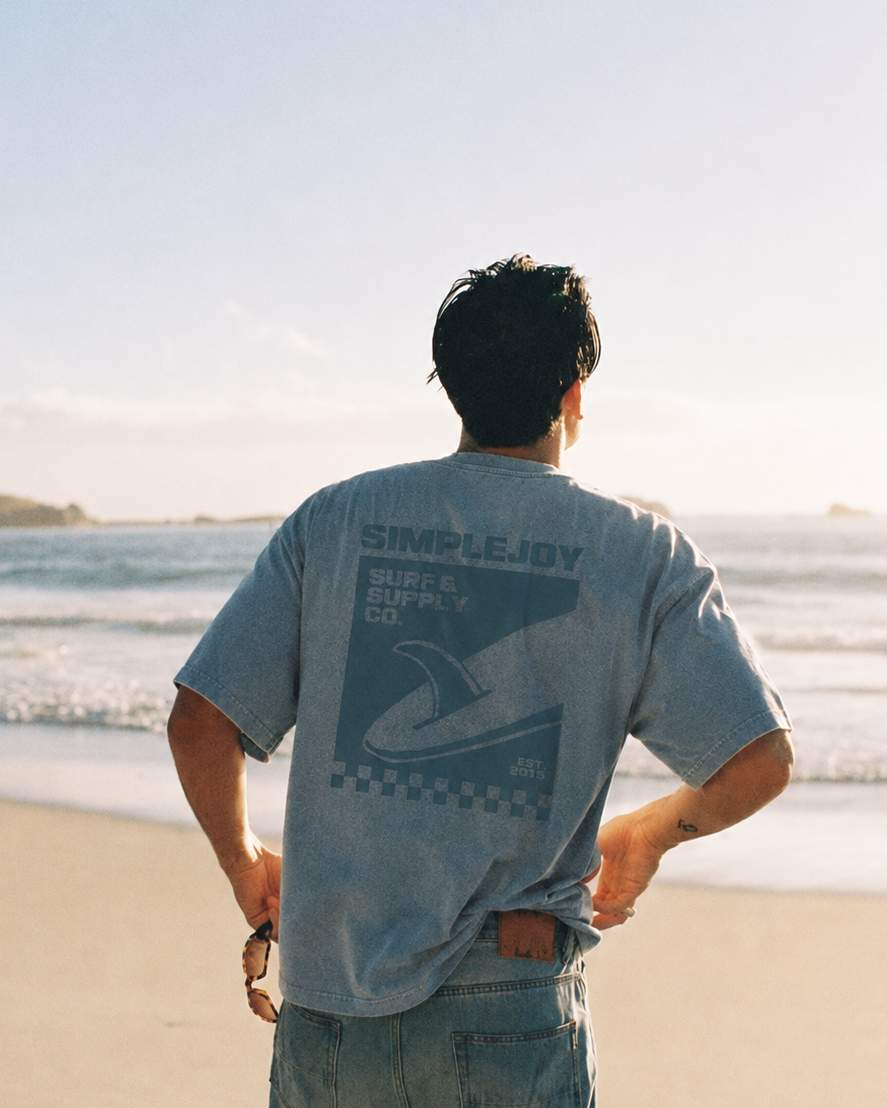 A man stands on a sandy beach facing the ocean, viewed from behind. He is wearing a light blue-gray t-shirt with a dark graphic print on the back that reads 'SIMPLEJODY SURF SHOP CO.
