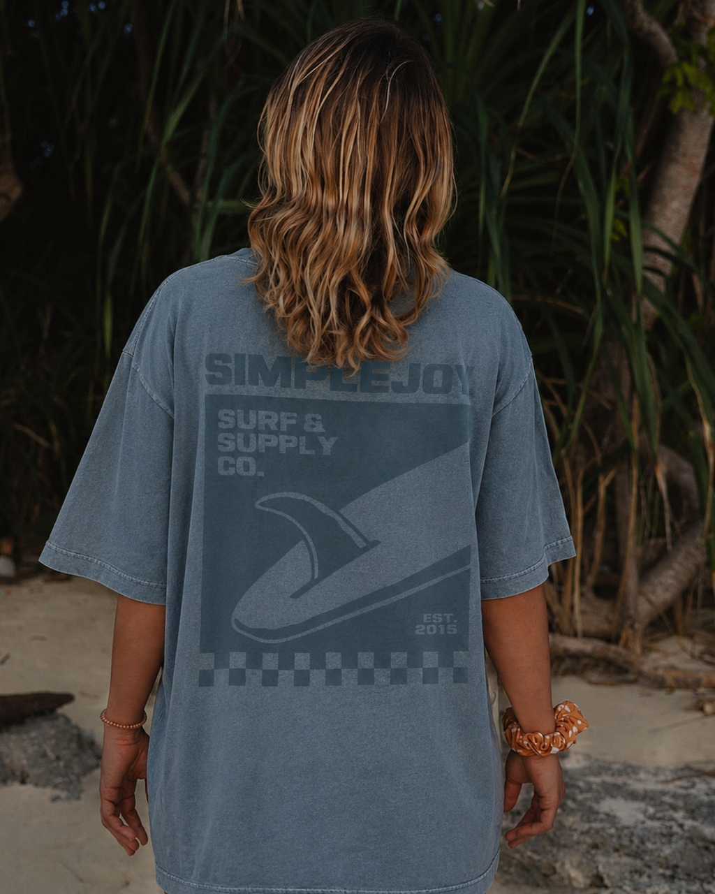 A person viewed from behind wearing a faded blue-gray t-shirt with a large dark graphic on the back that reads 'SIMPLE JOY SURF & SUPPLY CO.' and shows a surfboard silhouette.