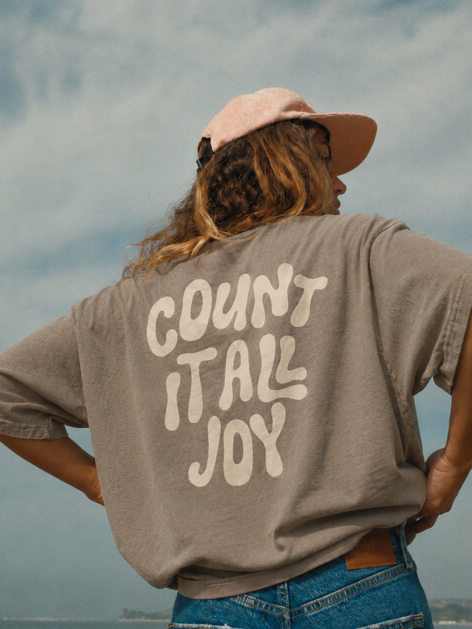 A person viewed from behind wearing a faded taupe-gray oversized t-shirt with 'COUNT IT ALL JOY' printed in cream across the back, paired with blue jeans and a light pink cap, set against a cloudy sky and water background.