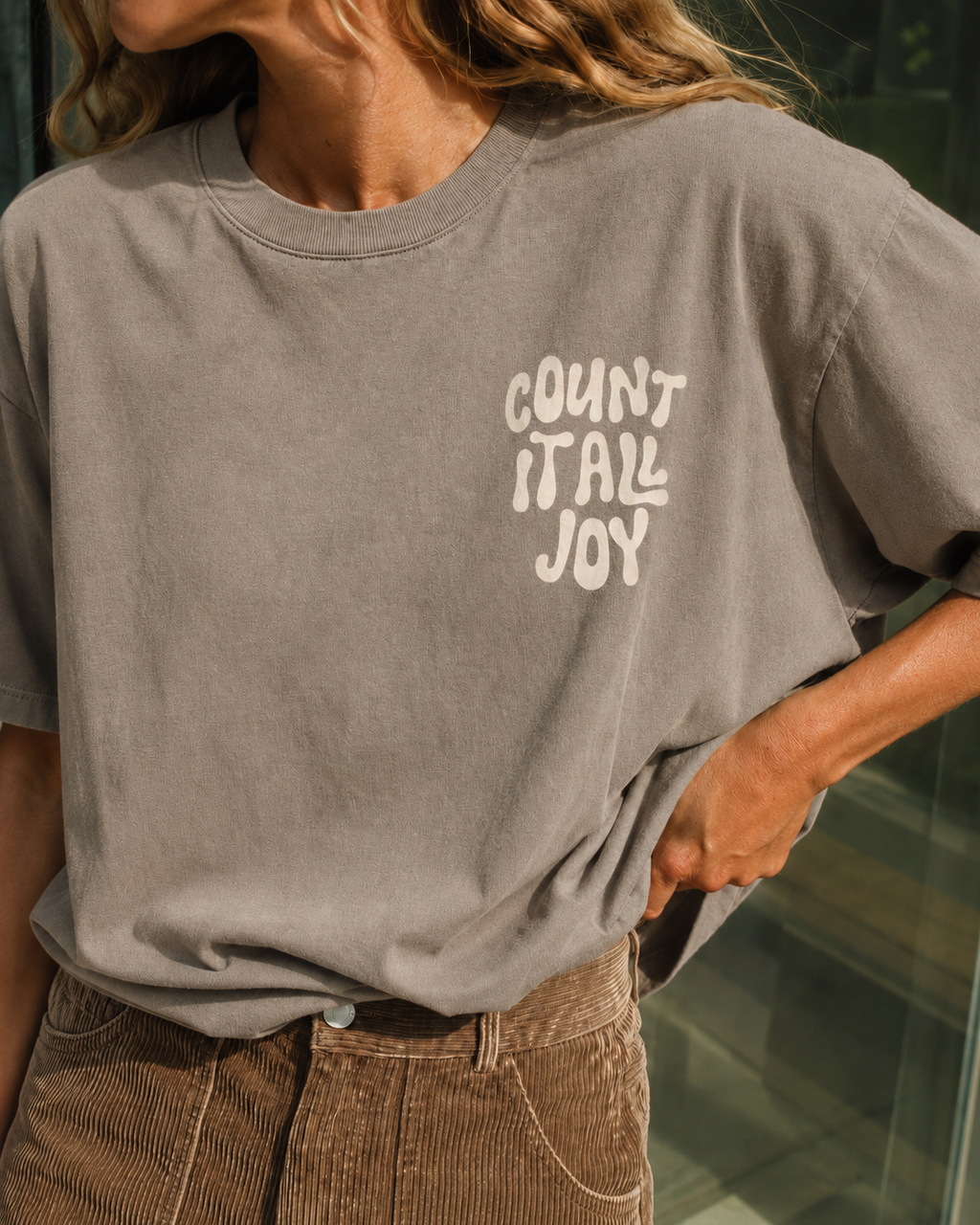 A close-up of a person wearing a washed taupe gray t-shirt with the phrase 'COUNT IT ALL JOY' printed in white on the chest, paired with brown corduroy pants.