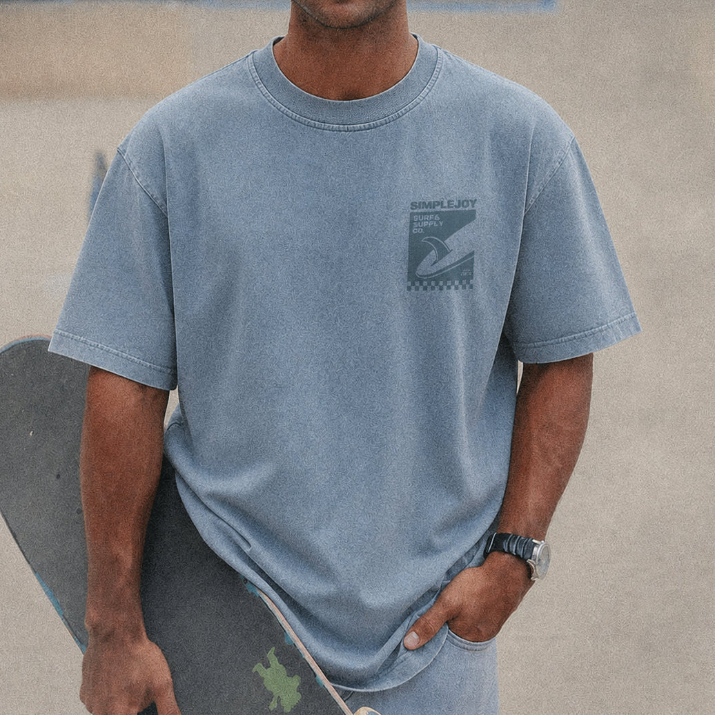 A man wearing a light blue crewneck t-shirt with a dark graphic on the left chest, holding a skateboard against a concrete background.