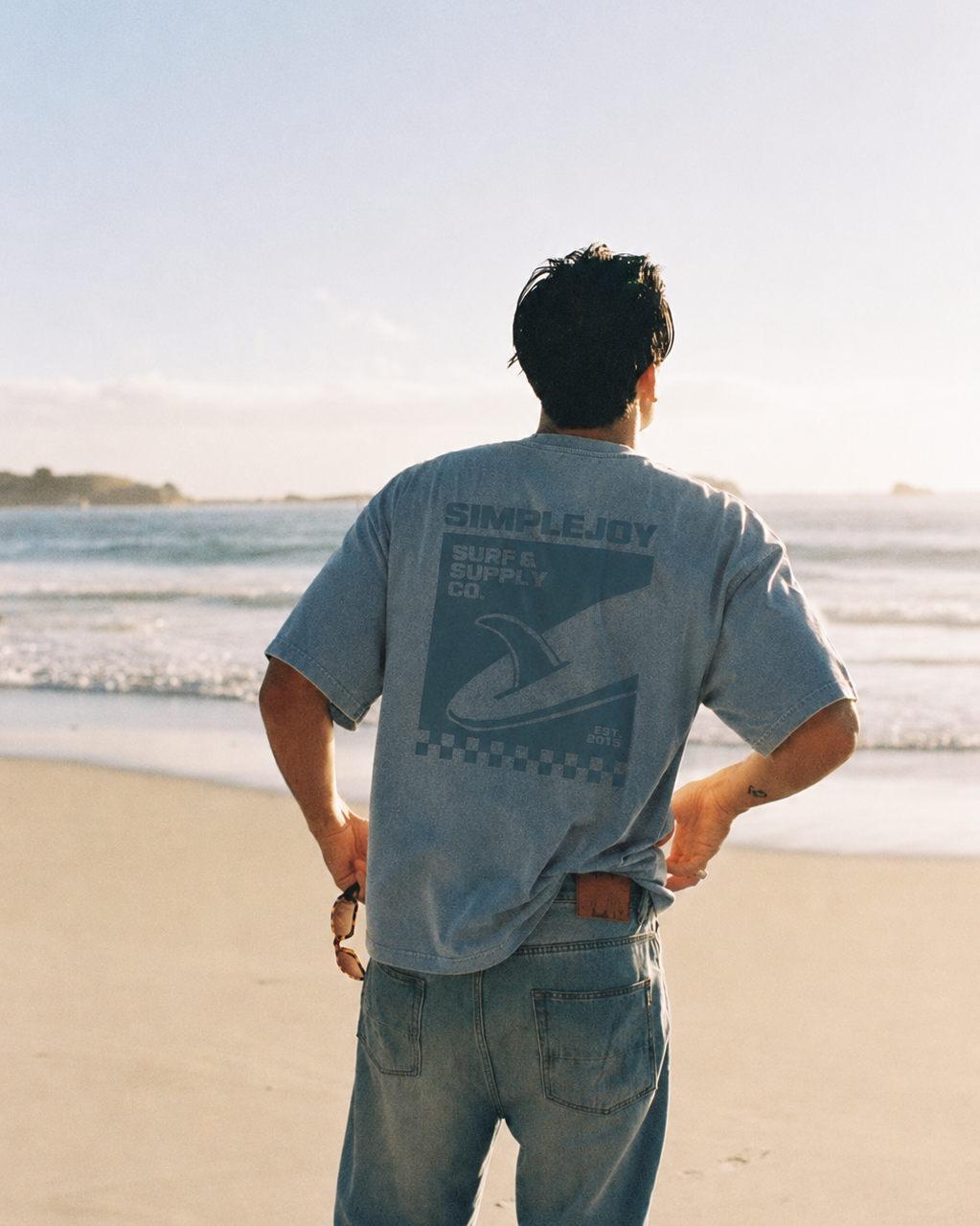 A man stands on a sandy beach with the ocean in the background, viewed from behind. He is wearing a light blue-gray t-shirt with a dark graphic print on the back that reads 'SIMPLEJODY SURF SHOP CO.