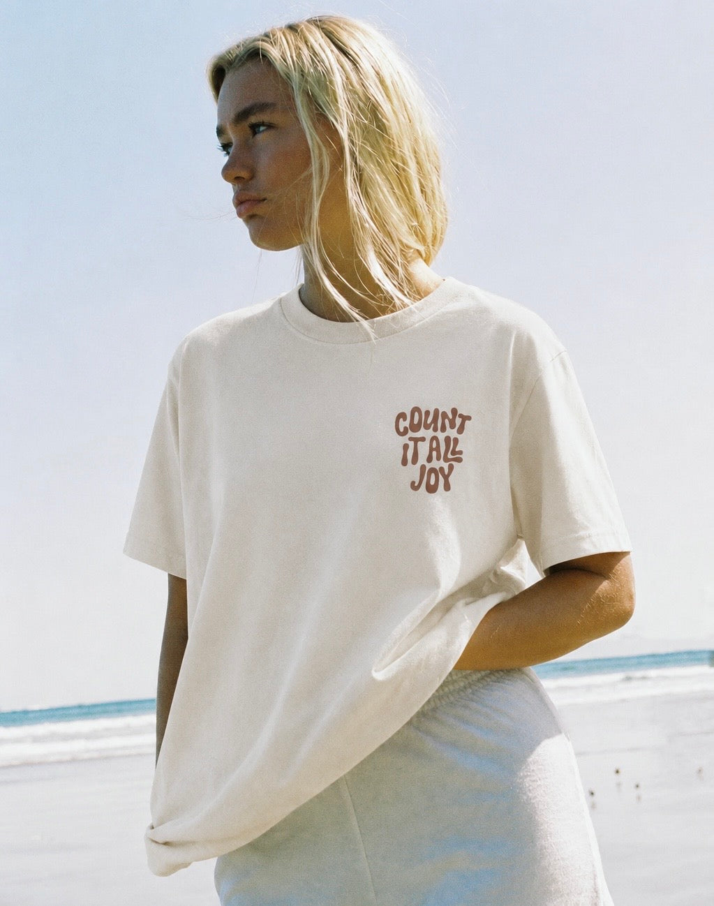 A woman wearing an oversized cream-colored t-shirt with the text 'COUNT IT ALL JOY' printed in a reddish-brown font on the left chest, set against a bright, sunlit beach background.