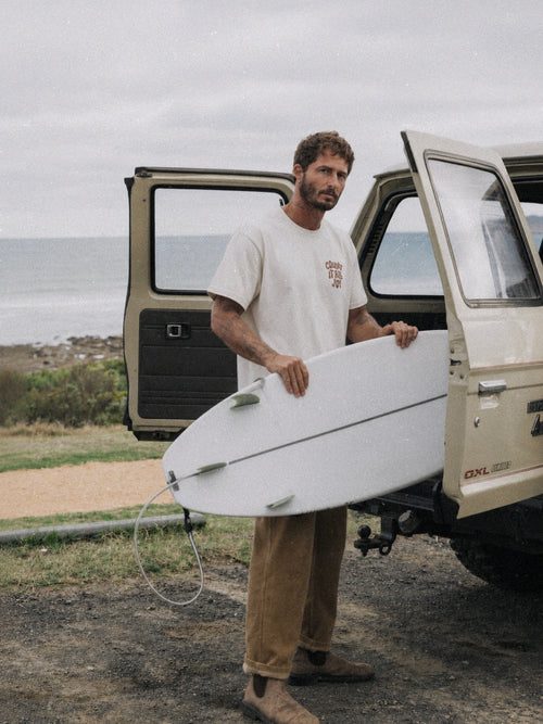 A man standing outdoors by an open beige vehicle, holding a white surfboard. He is wearing a cream graphic T-shirt, tan pants, and brown boots, with the ocean and cloudy sky in the background.